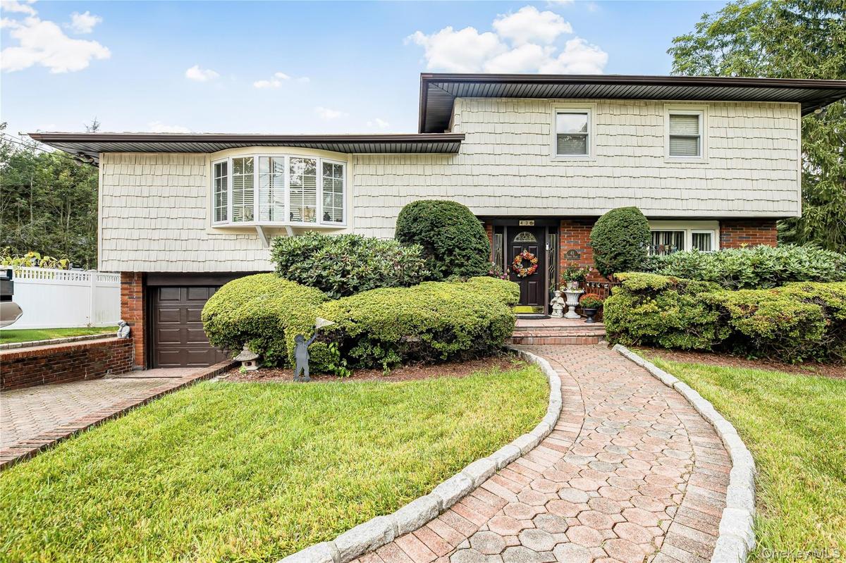 Split-level home featuring a brick paver walkway, mature shrubbery, a bay window, and a brown garage door