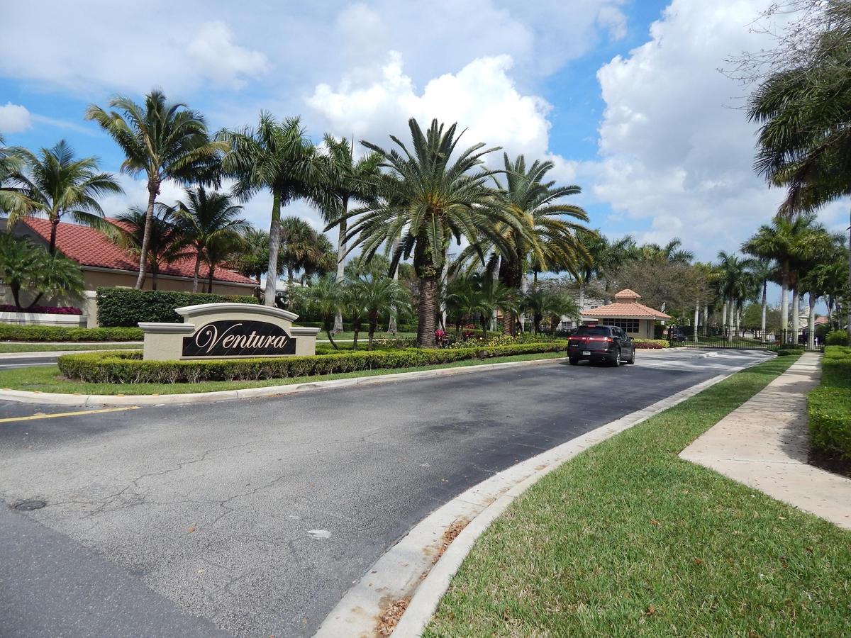Main entrance featuring a custom "Ventura" sign, lush tropical landscaping with mature palm trees, a dedicated entry road, and a security booth