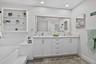 Dual basin vanity with white cabinetry and stone-finish countertop, large framed mirror, recessed lighting, built-in shelving, and a large soaking tub