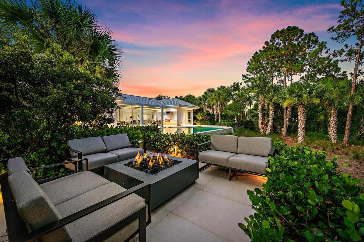 Paver patio with a built-in fire pit, lush landscaping, and mature palm trees