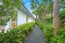 Gravel pathway bordered by abundant green shrubbery, mature trees, and a white exterior wall with multiple windows
