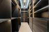 Custom gentleman’s walk-in wardrobe featuring dark wood cabinetry, extensive shelving and hanging space, integrated drawers, and sophisticated lightin