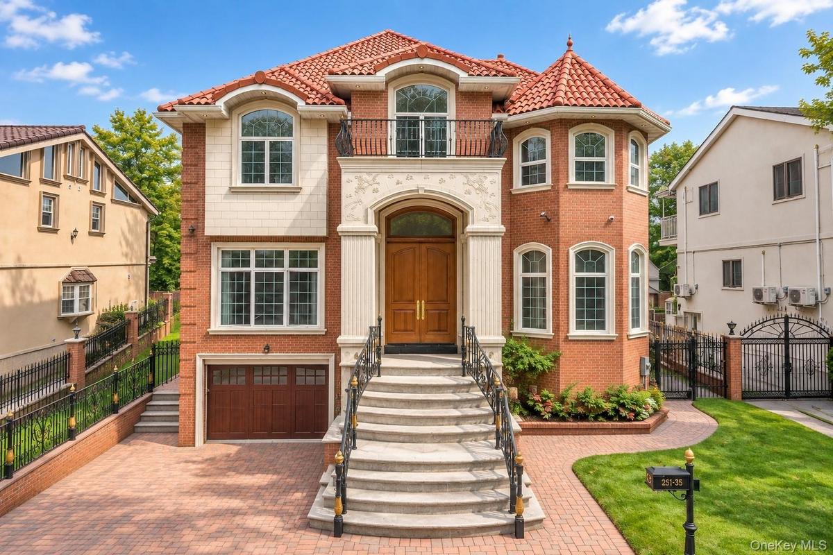 Red brick exterior with a terracotta tile roof, prominent arched entryway, a stone balustrade balcony, and a paved driveway with a garage door