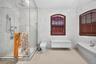 Glass-enclosed shower with multiple showerheads, white tile walls, two arched windows with wood-tone blinds, and a built-in bathtub with a white count