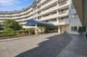 Curved multi-story building exterior with individual balconies, covered entryway with a blue canopy, and a paver driveway