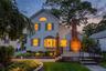 White shingle siding with gray shutters, bay window, prominent brick chimney, multi-level patio with black railings, and stone-edged landscaping with 