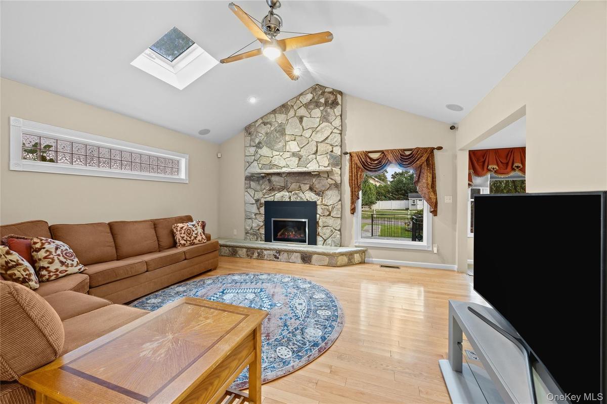 Vaulted ceiling with skylight, stone-clad fireplace, wood-finish flooring, and a ceiling fan