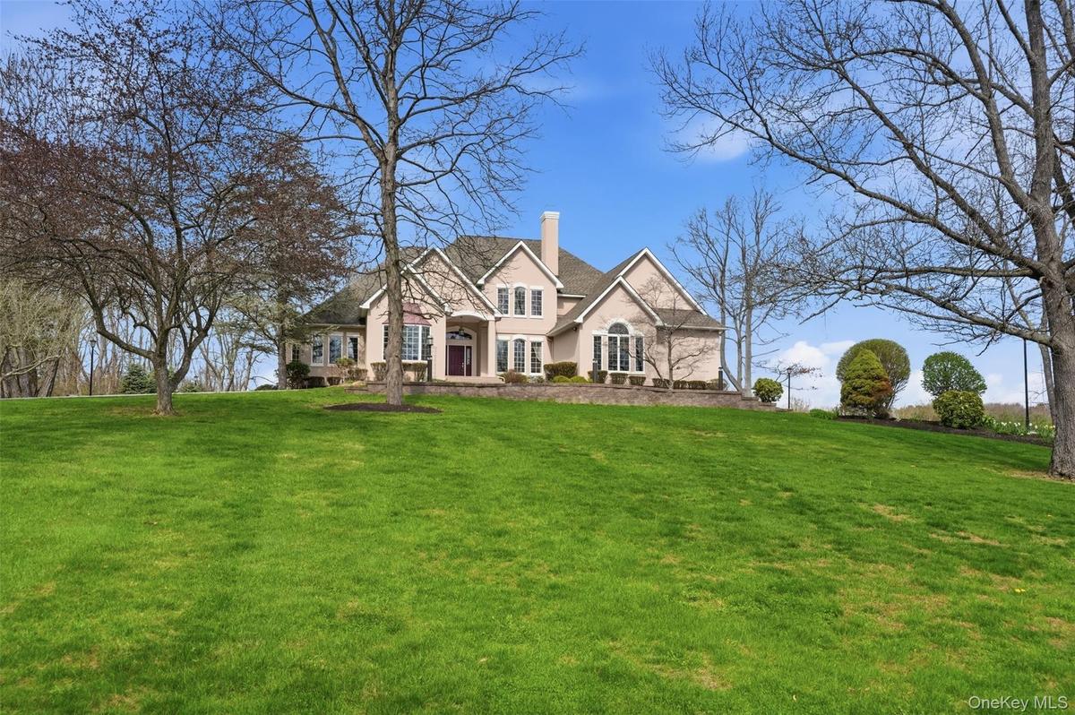 The property features a spacious front lawn with mature trees and a manicured landscape, set against a clear blue sky