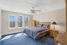 This bright bedroom features a ceiling fan, plush blue carpet, and a trio of windows offering views of the outdoors