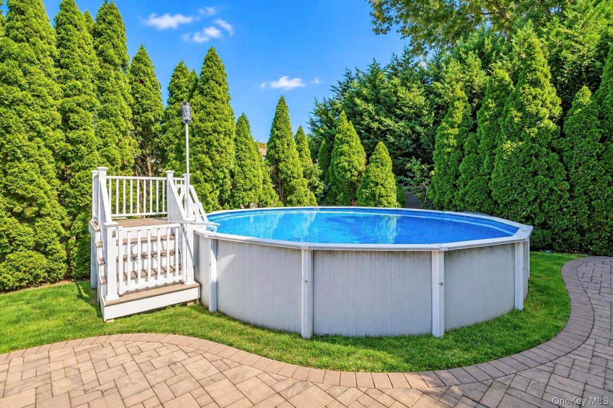 Above-ground pool with surrounding foliage, white wood-finish pool deck, and paver patio