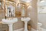Dual pedestal sinks with brass fixtures, gold-framed decorative mirrors, and wall-mounted sconces