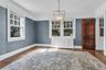 Hardwood flooring with a matte finish, blue textured wall covering, a dark wood panel door, white window casings, and a geometric chandelier