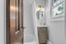 White subway tile shower surround, single window with white trim, gray vanity with integrated sink, gold-toned faucet, and oval wall mirror