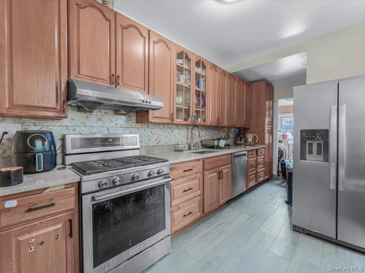 Kitchen with stainless steel appliances, under cabinet range hood, backsplash, glass insert cabinets, and light stone countertops