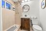 Walk-in shower with tiled walls, natural light window, dark wood-finish vanity with countertop sink, and a circular wall-mounted mirror