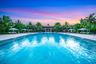 Expansive outdoor pool with a surrounding stone deck, tropical landscaping, and a distant clubhouse structure