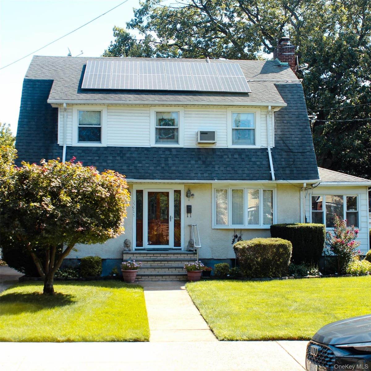 Gambrel roofline with solar panels, white siding, brick chimney, covered entry, and bay window