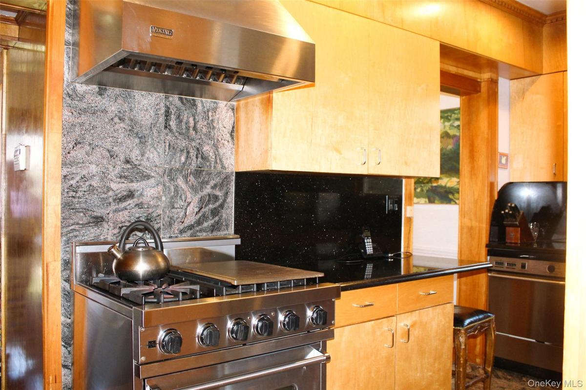 Stainless steel range with a professional-grade hood, natural wood-finish cabinetry, black stone countertops, and a tiled stone backsplash