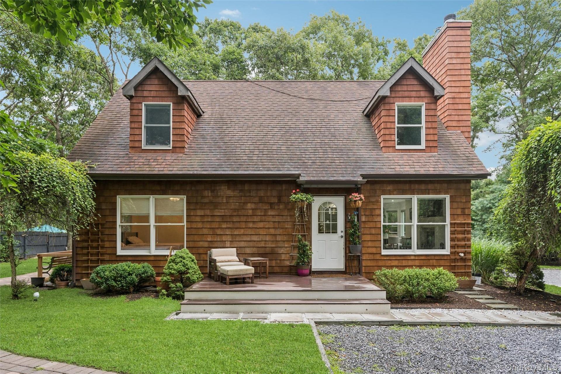 Cedar shake siding, dormer windows, a prominent brick chimney, a front deck, and mature landscaping