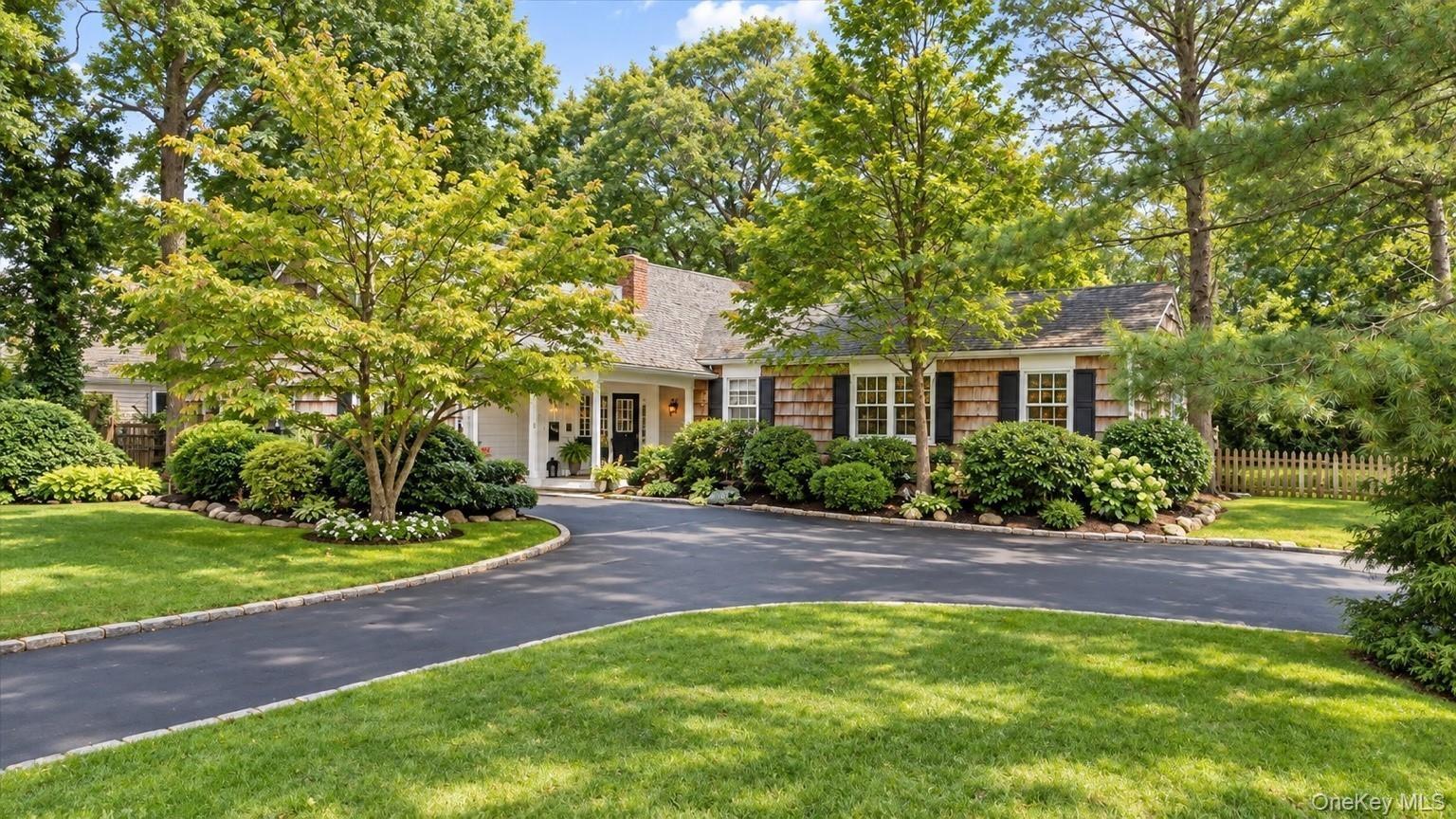 Asphalt driveway, manicured lawn, shaker siding, mature trees, and a covered entry