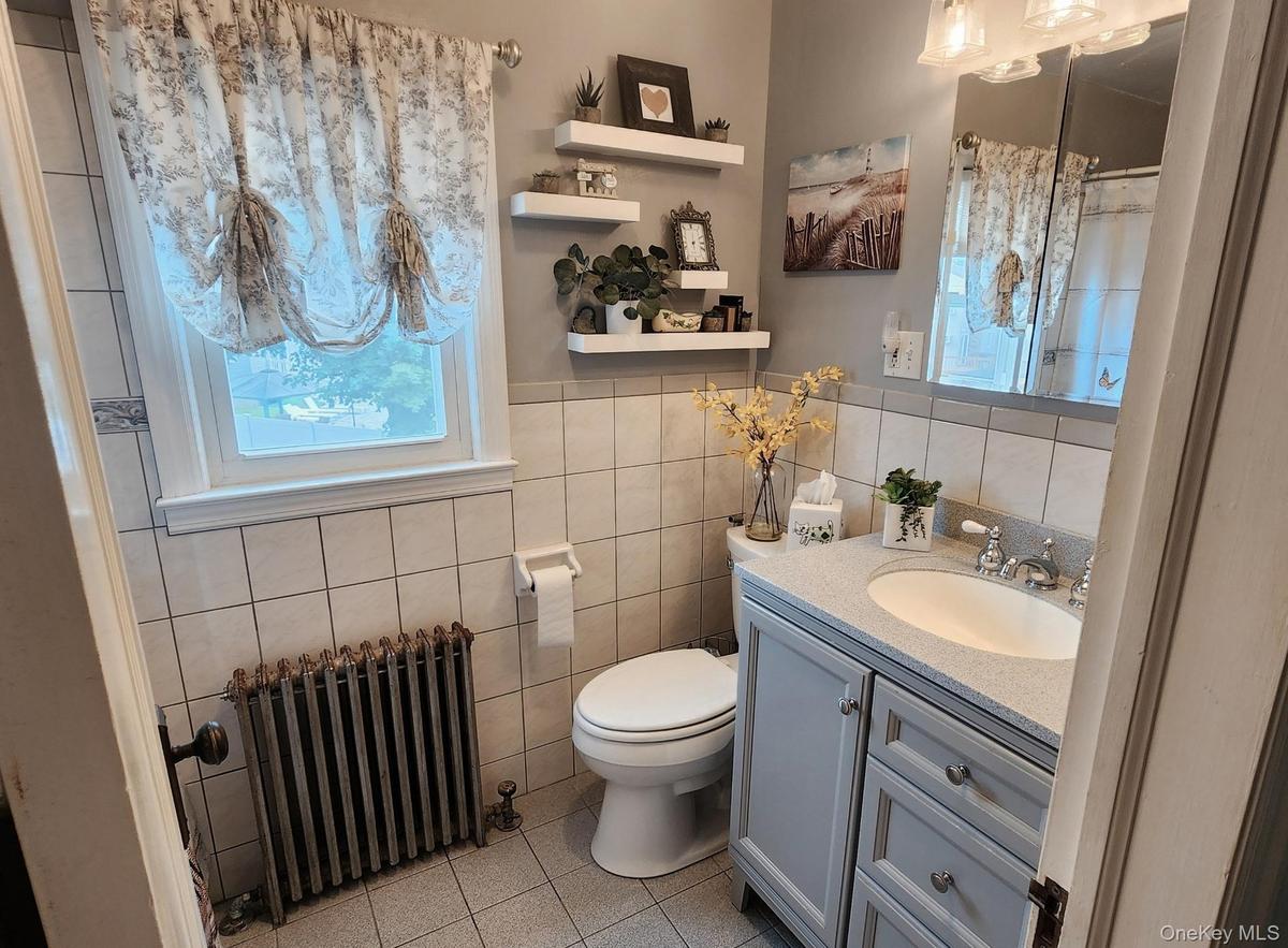 Bathroom featuring a window, pedestal sink with vanity, wall-mounted mirror, and tiled walls
