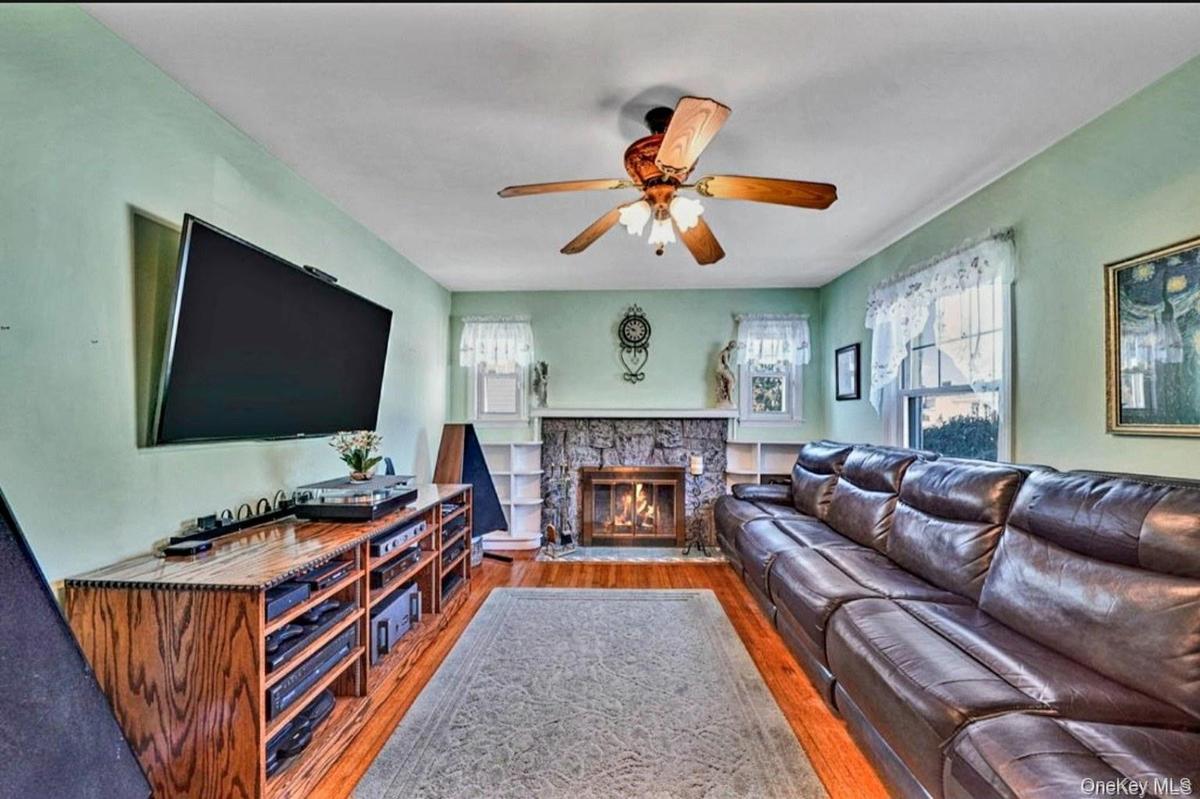 Hardwood flooring complemented by a stone-faced wood burning fireplace, a ceiling fan with integrated lighting, and multiple windows