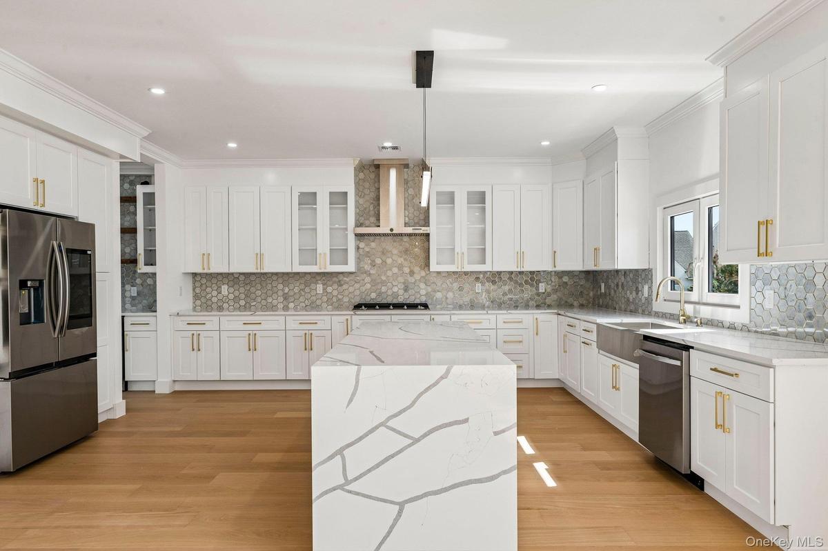 Gourmet kitchen featuring white shaker cabinetry, stainless steel appliances, a waterfall island, hexagon tile backsplash, and wood-finish flooring