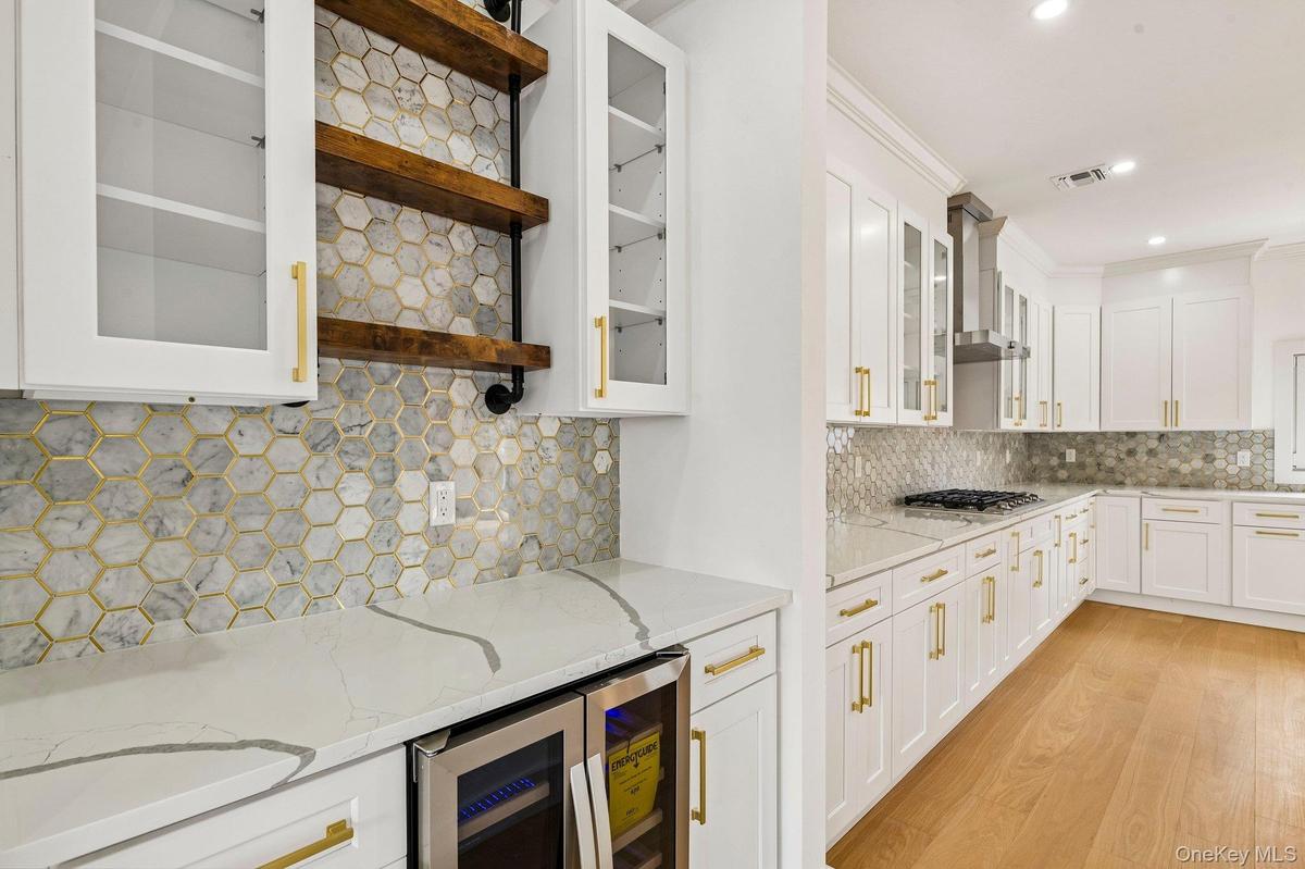 White cabinetry with gold-tone hardware, hexagon tile backsplash, quartz countertops, built-in beverage refrigerator, and wood-finish flooring