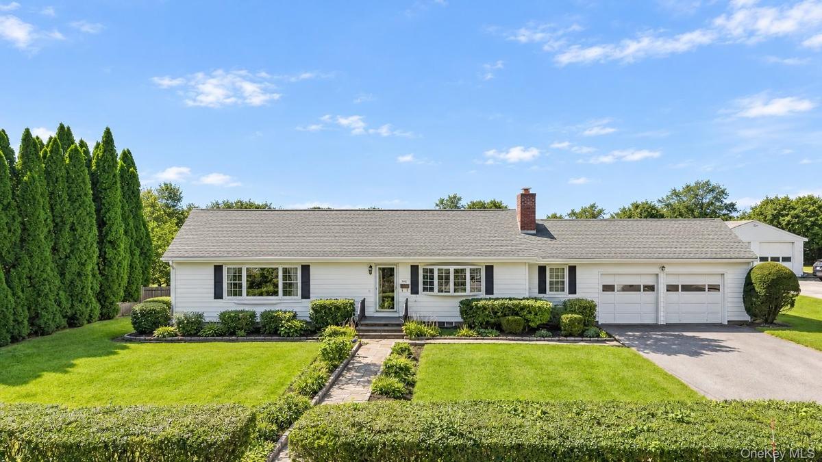 White ranch-style residence featuring a two-car garage, brick chimney, established front landscaping, and a paved driveway