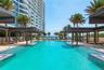 Expansive swimming pool with integrated steps, surrounded by lounging areas under pergolas, palm trees, and a multi-story building