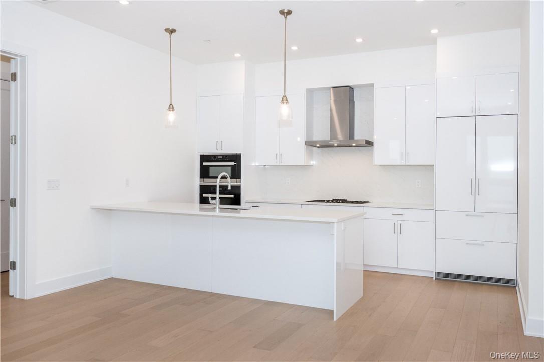 Kitchen featuring stainless steel double oven, wall chimney range hood, white cabinetry, light wood-type flooring, and decorative light fixtures