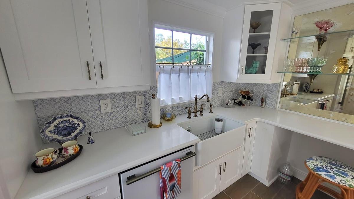 White shaker-style cabinetry, patterned tile backsplash, farmhouse-style sink with a brass-finish faucet, and a built-in dishwasher