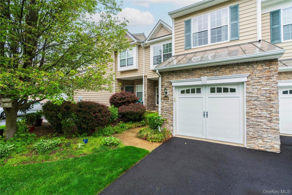Exterior featuring stacked stone accents, vinyl siding, a two-car garage with paneling and windows, a glass canopy, and established landscaping