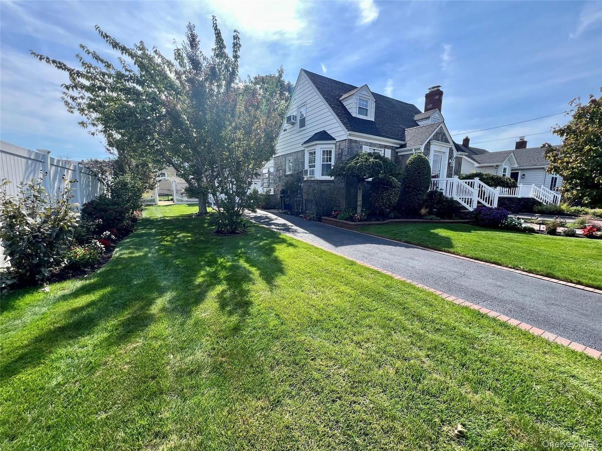 Gable roofline, bay window, white picket fence, paved driveway with brick edging, and mature trees on a lush lawn
