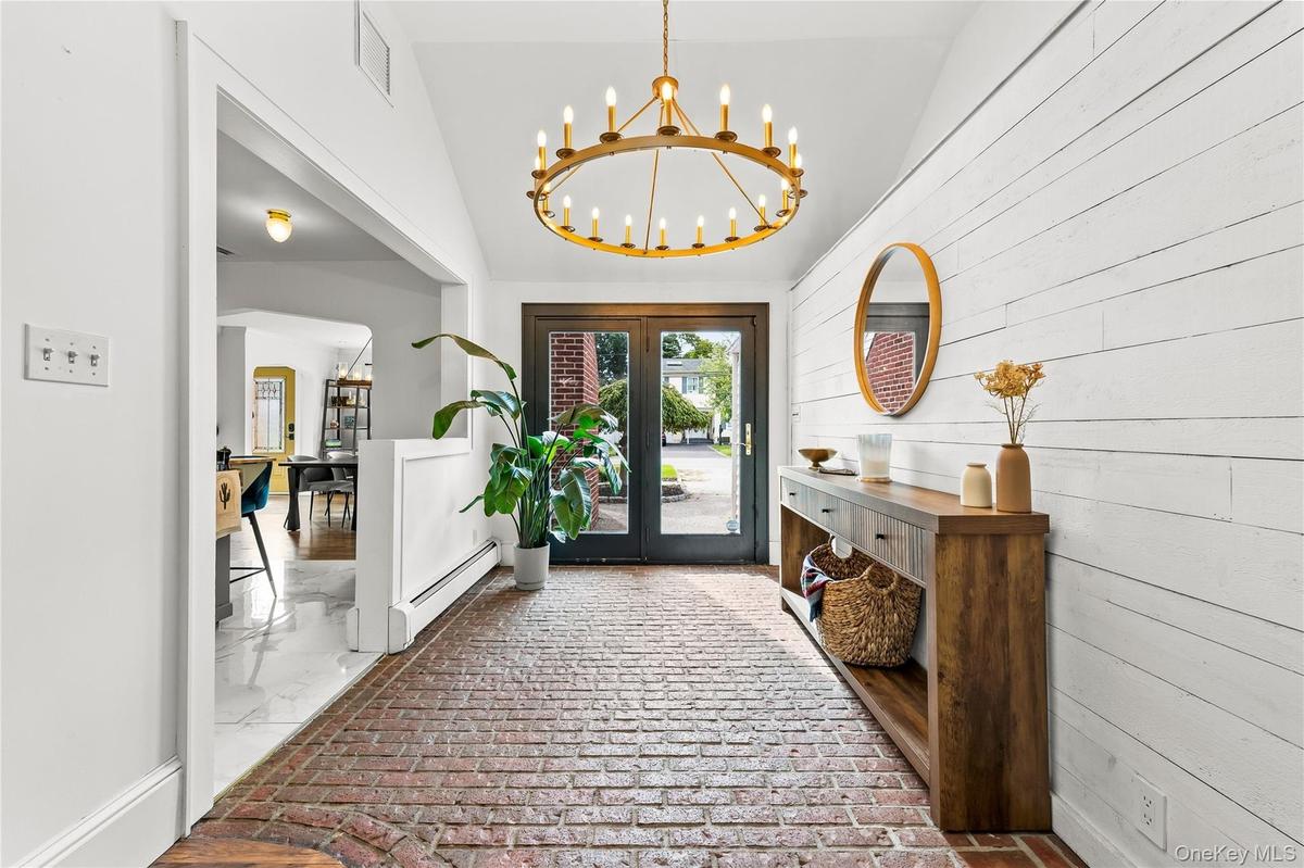Brick flooring, vaulted white ceiling, shiplap wall, brass chandelier, and double entry doors