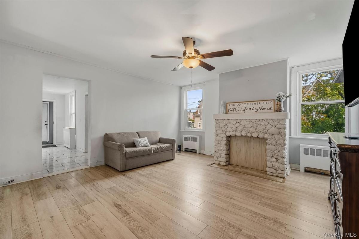 Light wood-finish flooring, stone-clad fireplace with mantel, ceiling fan with integrated lighting, and multiple windows