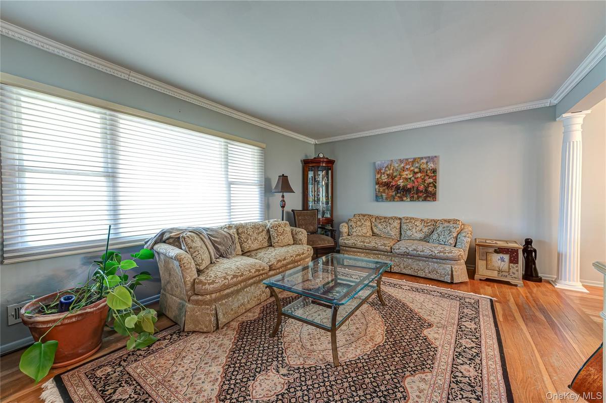 Hardwood flooring, crown molding, a large picture window with blinds, and decorative fluted columns