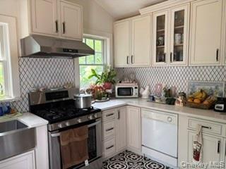 White cabinetry with glass-front accents, stainless steel range with hood, patterned tile backsplash, and farmhouse sink