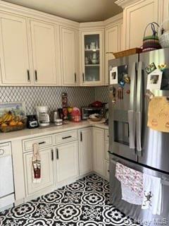 White cabinetry with crown molding, glass-front corner cabinet, stainless steel refrigerator, patterned floor tile, and light-toned countertops