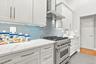 White cabinetry with brushed nickel hardware, quartz countertops, a stainless-steel range with a ventilation hood, and a light blue tile backsplash