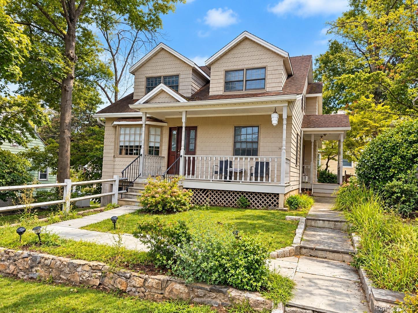Shingle siding exterior with dormer windows, covered front porch, stone retaining wall, paved walkway, and mature landscaping
