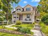 Shingle siding exterior with dormer windows, covered front porch, stone retaining wall, paved walkway, and mature landscaping