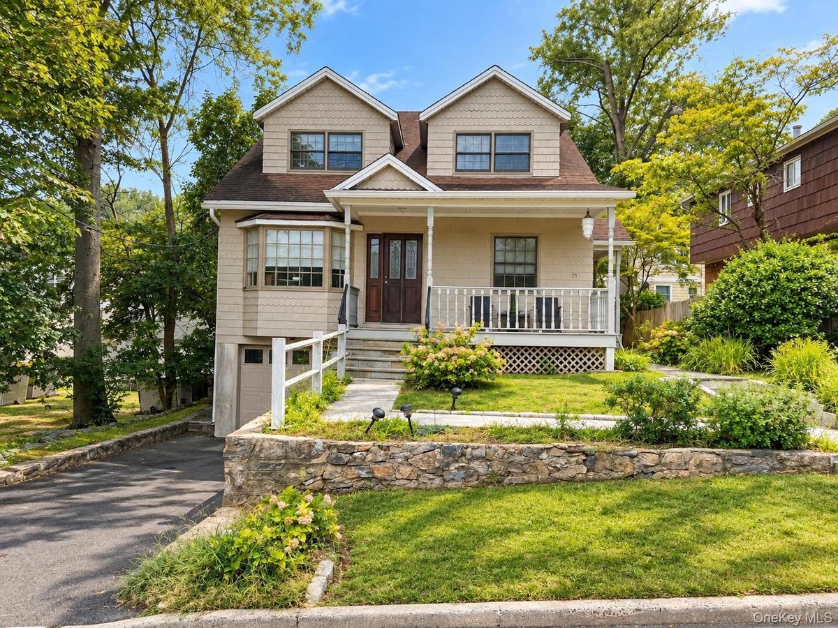 Dormer windows, covered front porch with white railing, stone retaining wall, paved driveway, and established landscaping
