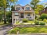 Dormer windows, covered front porch with white railing, stone retaining wall, paved driveway, and established landscaping
