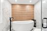 Freestanding soaking tub with a floor-mounted matte black faucet, wall-mounted wood-finish tile, and large format white tile with gold veining