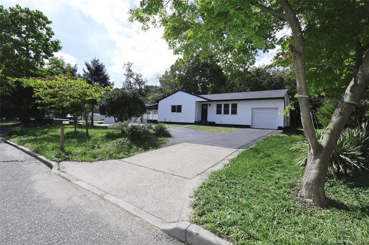 White siding exterior with a dark roofline, an attached garage, a paved driveway, and mature landscaping