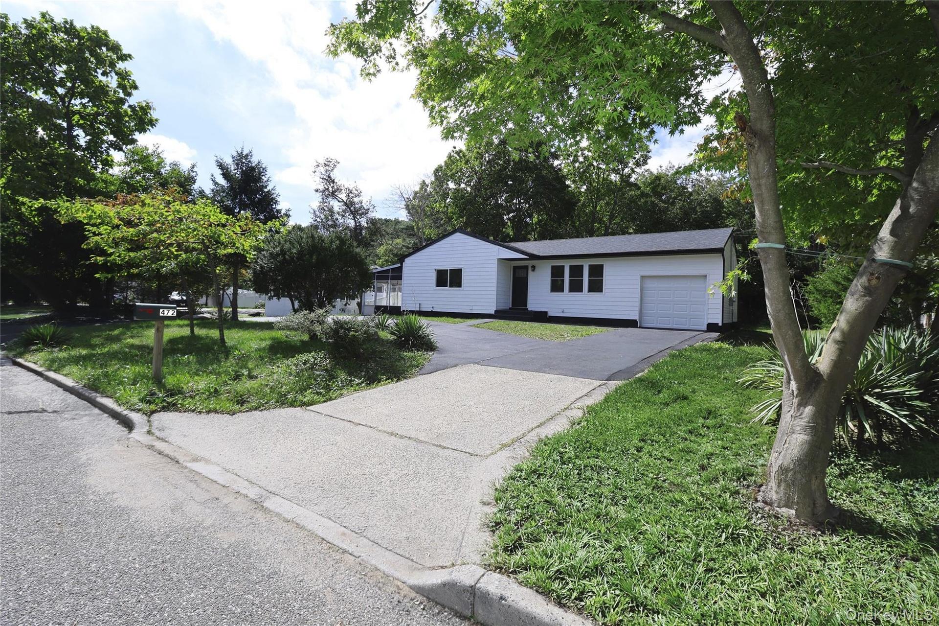White siding exterior with a dark roofline, an attached garage, a paved driveway, and mature landscaping