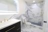 Vanity with integrated sink and gold-tone fixtures, backlit mirror, floor-to-ceiling marbled tile, and a walk-in shower with a recessed shelf
