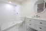 Bathroom featuring a white vanity with a stone countertop, a circular mirror, a walk-in shower with textured wall tiles, and glass corner shelves