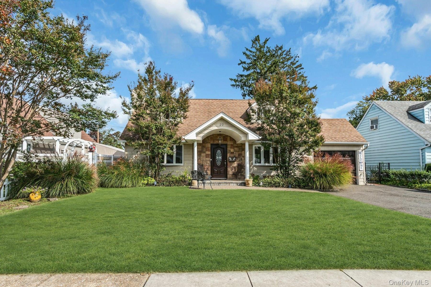 Pitched roof with asphalt shingles, stone facade entrance, attached garage with wood-finish door, paved driveway, and established landscaping